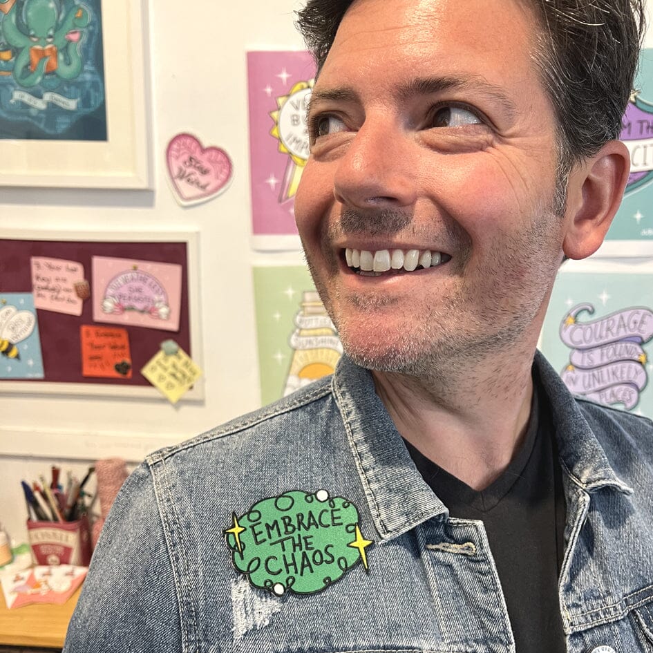 An iron on embroidered patch is being worn by a man on a denim jacket. The patch is green with yellow stars and reads Embrace The Chaos.