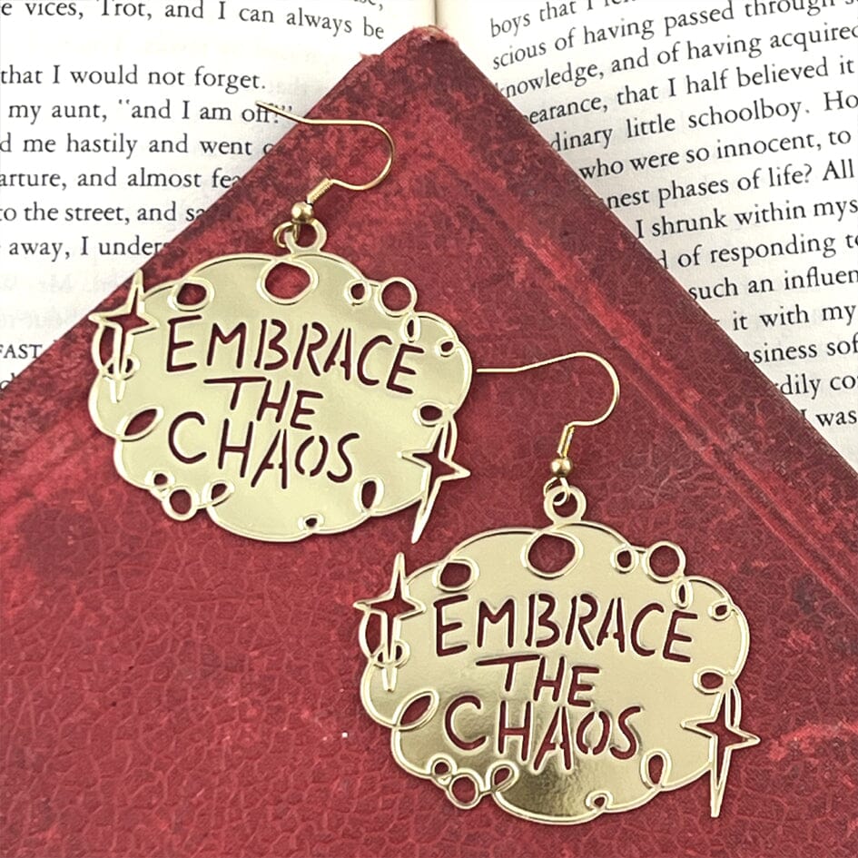 A pair of dangle brass earrings displayed on a red book background. The earrings read Embrace The Chaos.