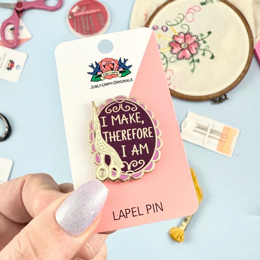 Lapel pin with 'I Make, Therefore I Am' design on a pink card held by a hand.
