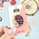 Lapel pin with 'I Make, Therefore I Am' design on a pink card held by a hand.