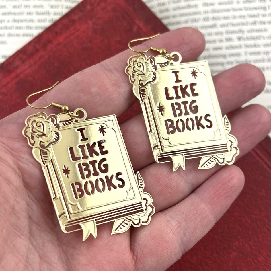 A pair of dangle brass earrings displayed on a hand. The earrings read I Like Big Books.
