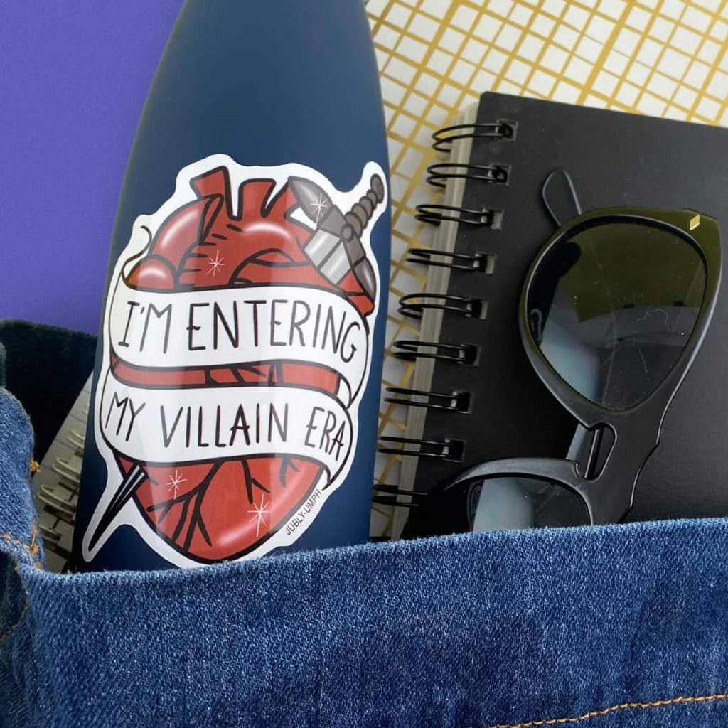 The vinyl sticker is in the shape of a human heart with a dagger. The sticker is on a blue water bottle. The sticker reads I'm Entering My Villain Era.