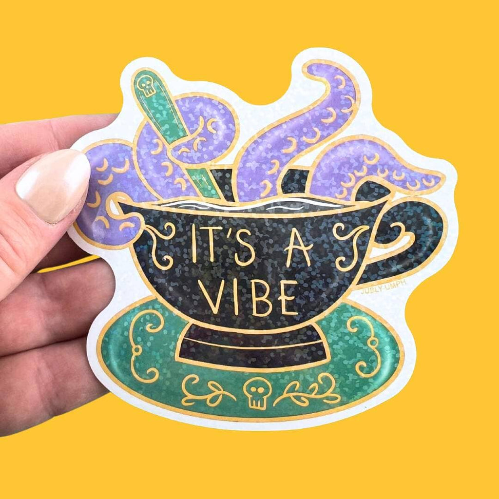 A vinyl sticker displayed in a hand on a yellow background. The sticker is in the shape of a black teacup with purple tenticles coming out. The pin reads It's A Vibe.