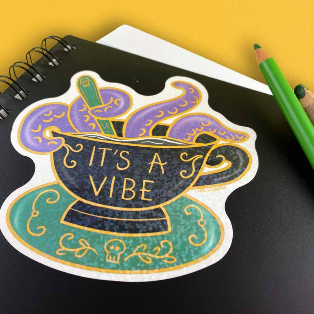 A vinyl sticker displayed on a black notebook. The sticker is in the shape of a black teacup with purple tenticles coming out. The pin reads It's A Vibe.