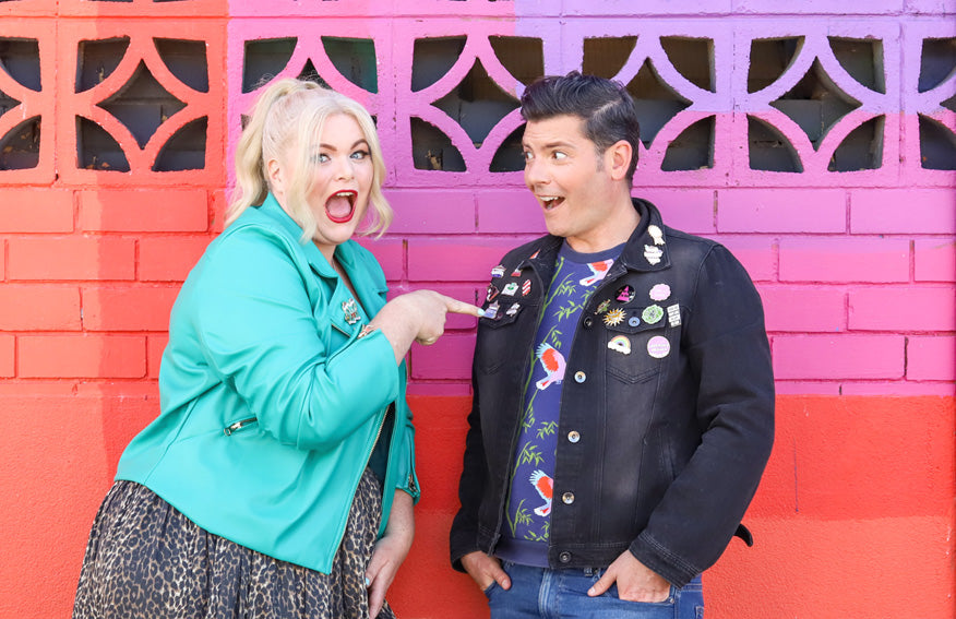 Two people standing in front on a brightly coloured wall wearing denim jackets covered in funny and empowering enamel lapel pins. 