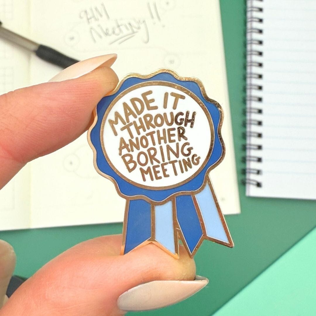 Enamel pin with 'Made it through another boring meeting' text held by a hand.