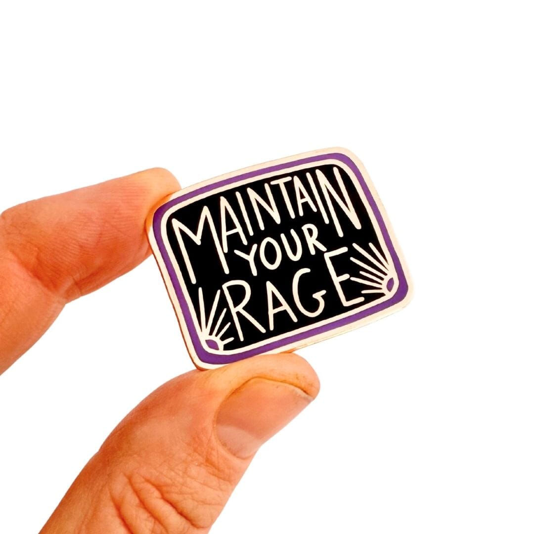 Enamel pin with 'Maintain Your Rage' text held between fingers on a white background