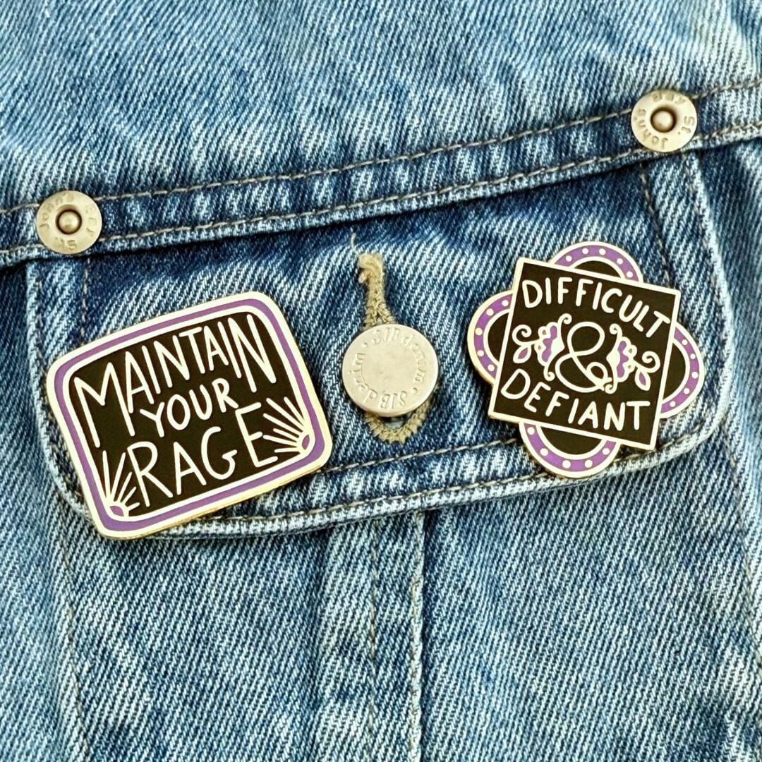Two enamel pins on a denim jacket, one reading 'Maintain Your Rage' and the other 'Difficult & Defiant'.