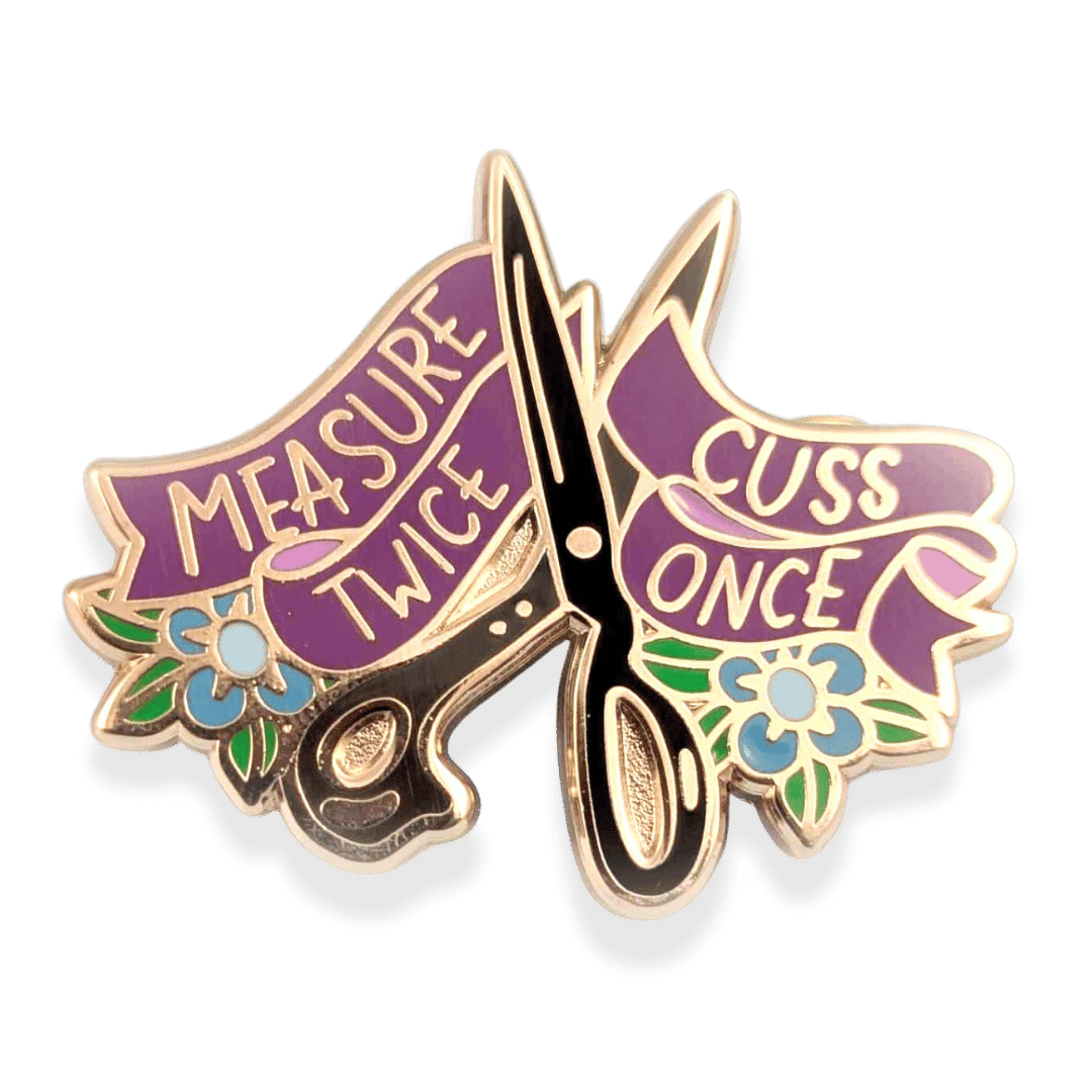 Scissor-shaped pin with 'Measure Twice, Cuss Once' text with blue flowers and green foliage.