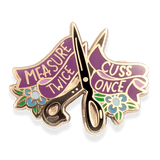 Scissor-shaped pin with 'Measure Twice, Cuss Once' text with blue flowers and green foliage.