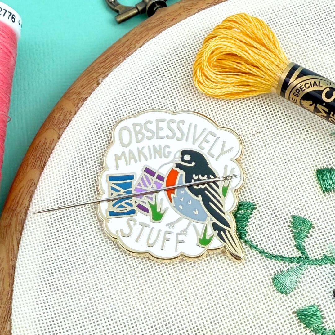 Embroidery hoop with a bird-themed enamel pin and yellow thread on a turquoise background