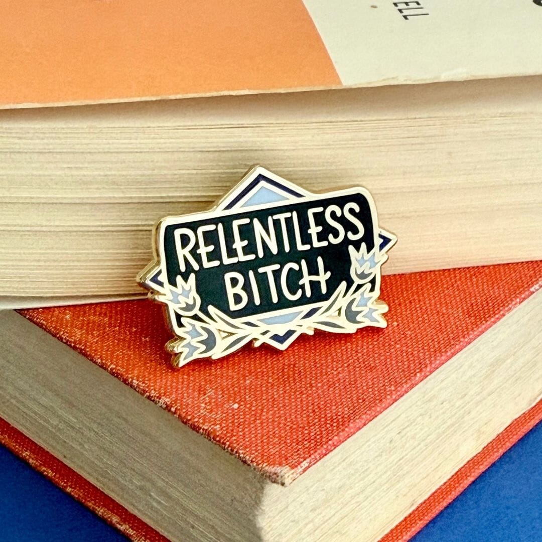 Pin with 'RELENTLESS BITCH' text on a stack of books
