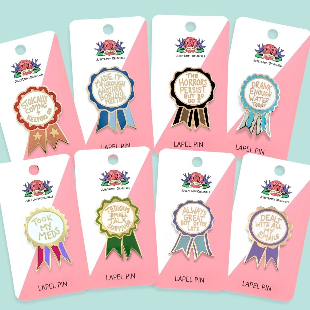 Set of eight lapel pins on pink cards with colorful ribbons and text, against a light blue background.