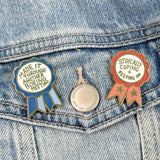 Two pins on a denim jacket, one with a blue ribbon and text, the other with a red ribbon and text.