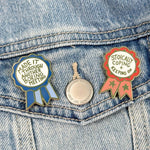 Two pins on a denim jacket, one with a blue ribbon and text, the other with a red ribbon and text.