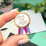 Pin with 'Took My Meds' text held by a hand on a desk with plants in the background