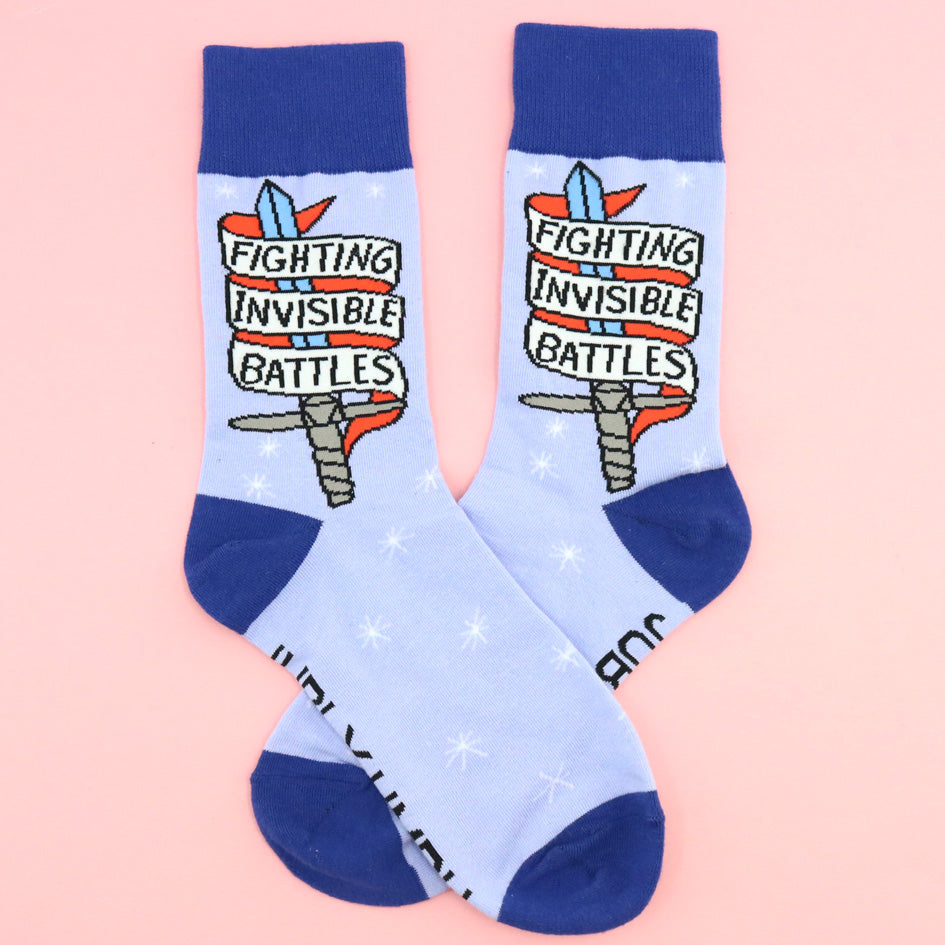 A pair of socks standing against a pink background. The socks are blue and read Fighting Invisible Battles inside a dagger.