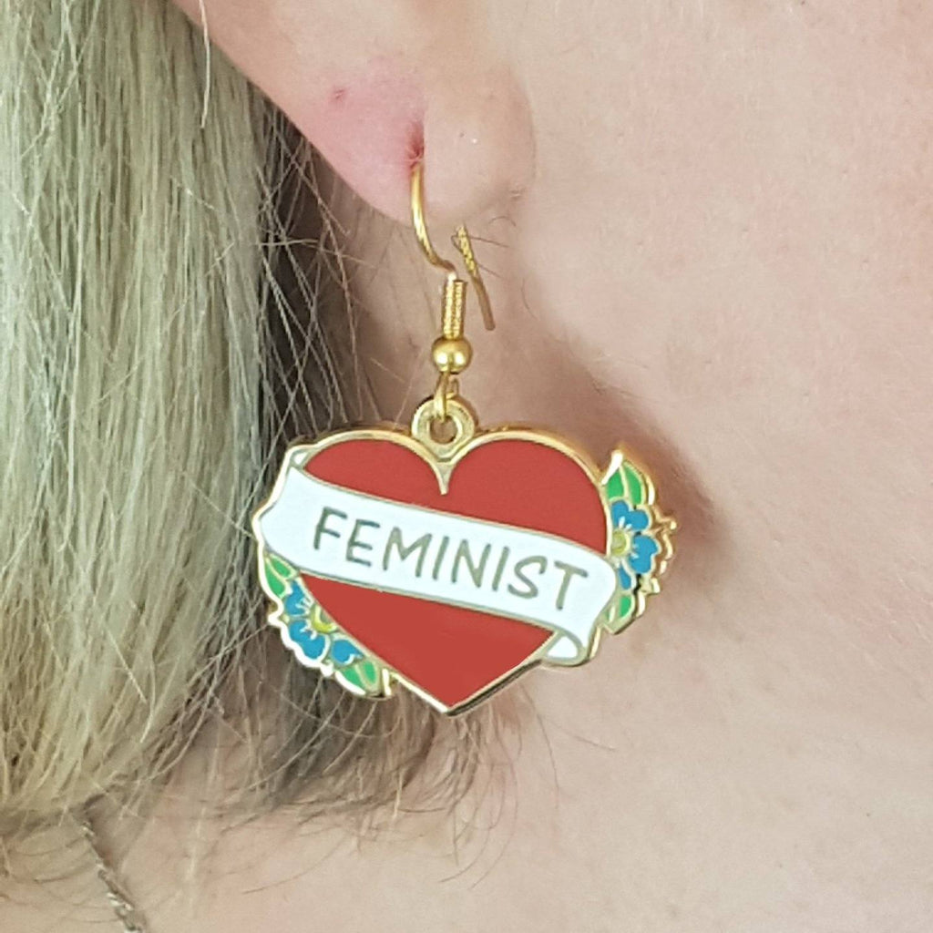 A single earring displayed from an ear lobe. The earrings are in the shape of a red heart and read Feminist.