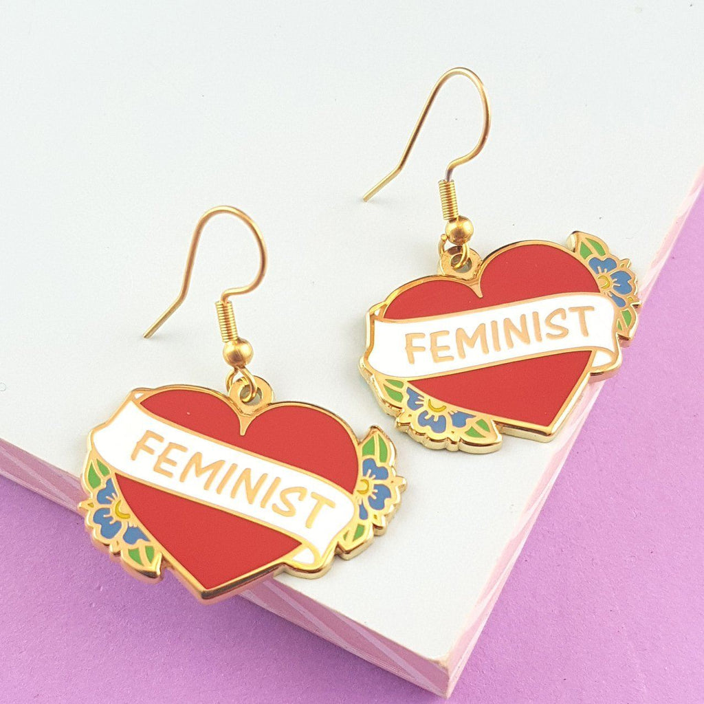 A pair of earrings displayed on a purple background. The earrings are in the shape of a red heart and read Feminist.
