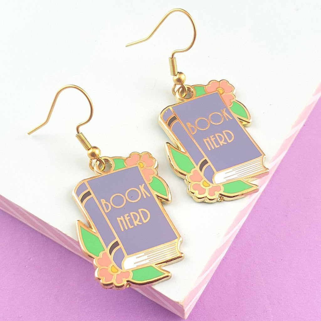 A pair of dangle earrings displayed on a white and pink background. The earrings say Book Nerd in the middle of a blue book.