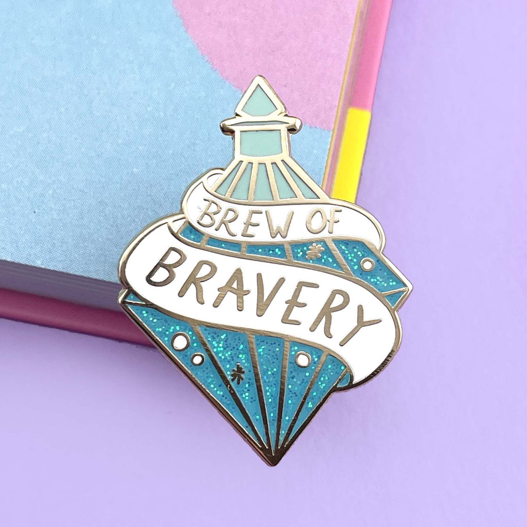 A hard enamel lapel pin on a purple background. The pin is in the profile of a diamond shaped bottle with blue glitter. The pin says Brew Of Bravery.