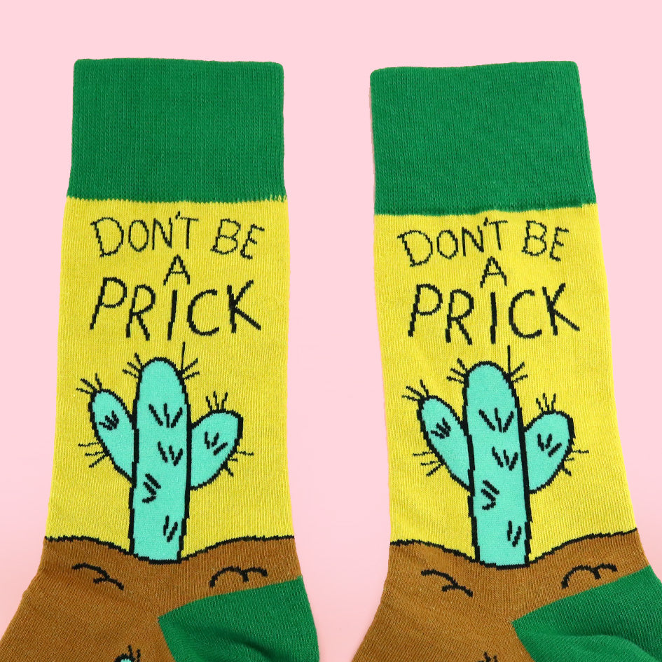 A pair of socks against a pink background. The socks are yellow, green and brown with a teal cactus. The socks read Don't Be A Prick.