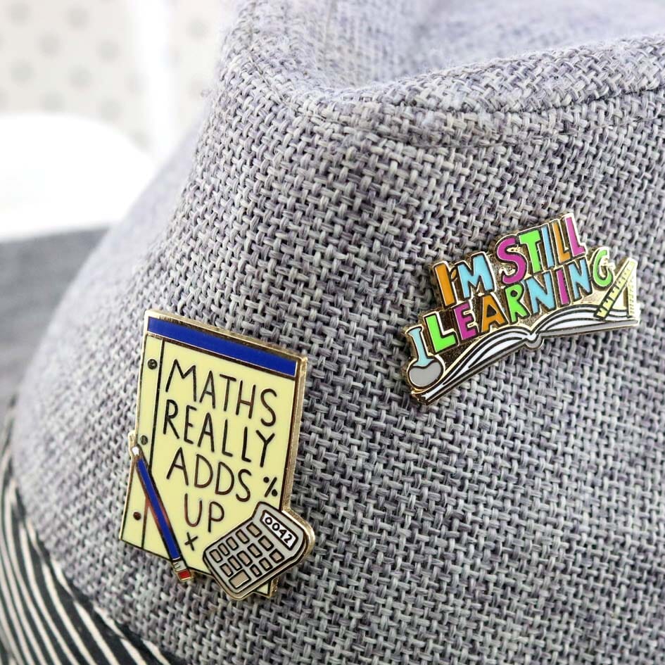 Two hard enamel pins being displayed on a gray hat. The first lapel pin is an open book and reads I'm Still Learning. The second lapel pin is reads Maths Really Adds Up.