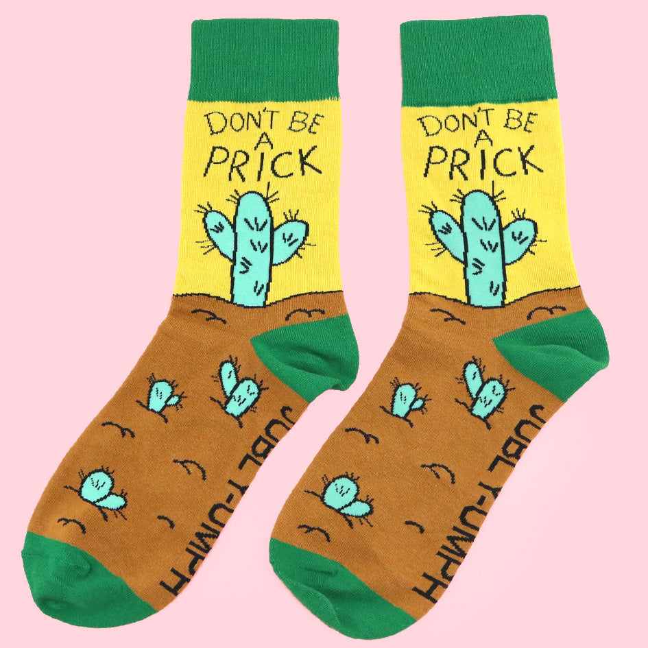 A pair of socks against a pink background. The socks are yellow, green and brown with a teal cactus. The socks read Don't Be A Prick.