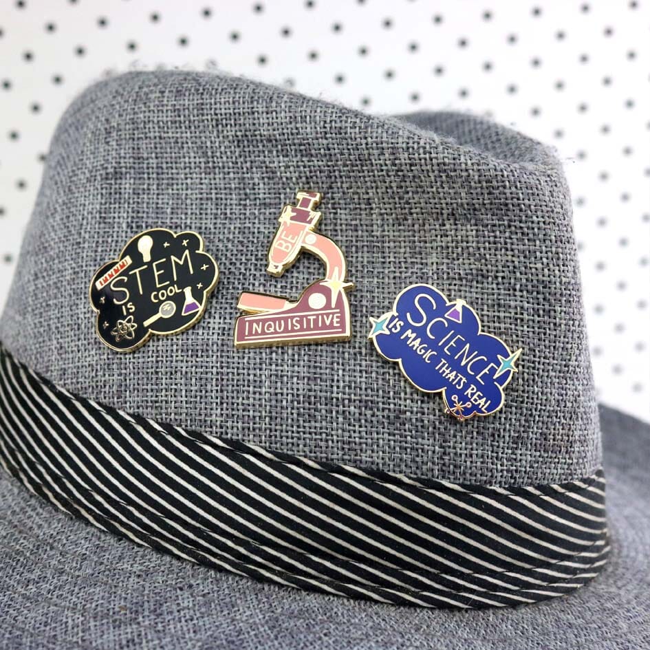 A hard enamel lapel pin on a gray hat. The pin is in the shape of a microscope and reads Be Inquisitive. There are two more lapel pins reading Science is Magic That’s Real and STEM is Cool.