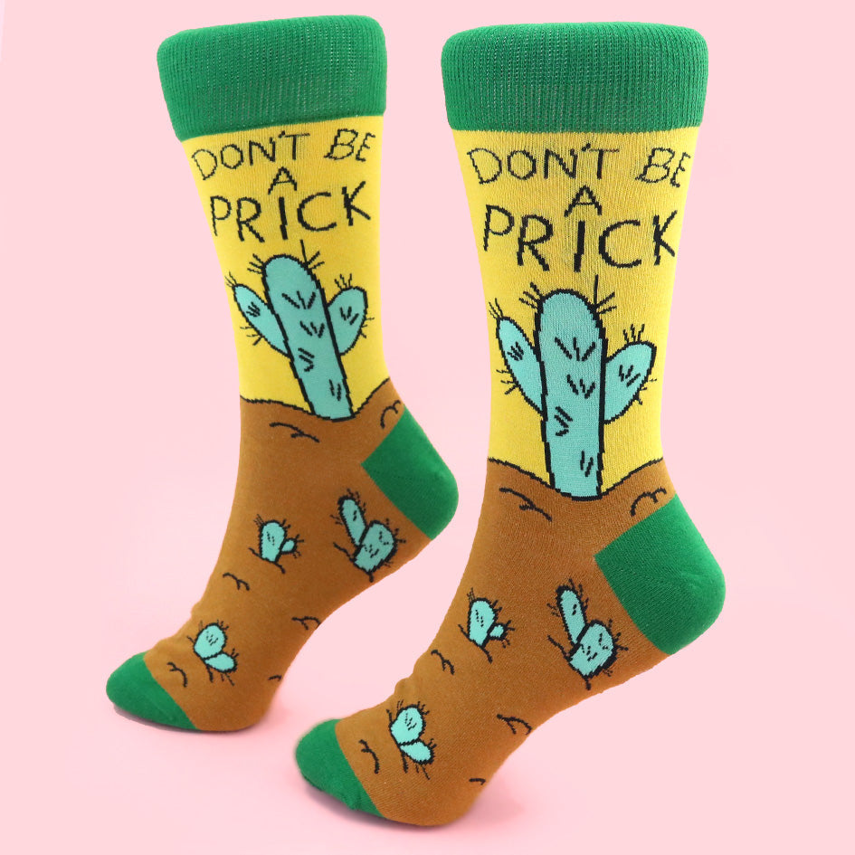 A pair of socks standing against a pink background. The socks are yellow, green and brown with a teal cactus. The socks read Don't Be A Prick.