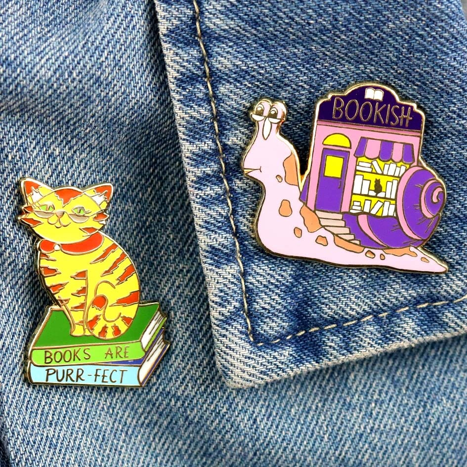 Two hard enamel lapel pins on a denim jacket. The first pin says Books Are Purr-fect with a cat sitting on books. The second pin says Bookish. The pin design is a snail with a bookshop on its back.