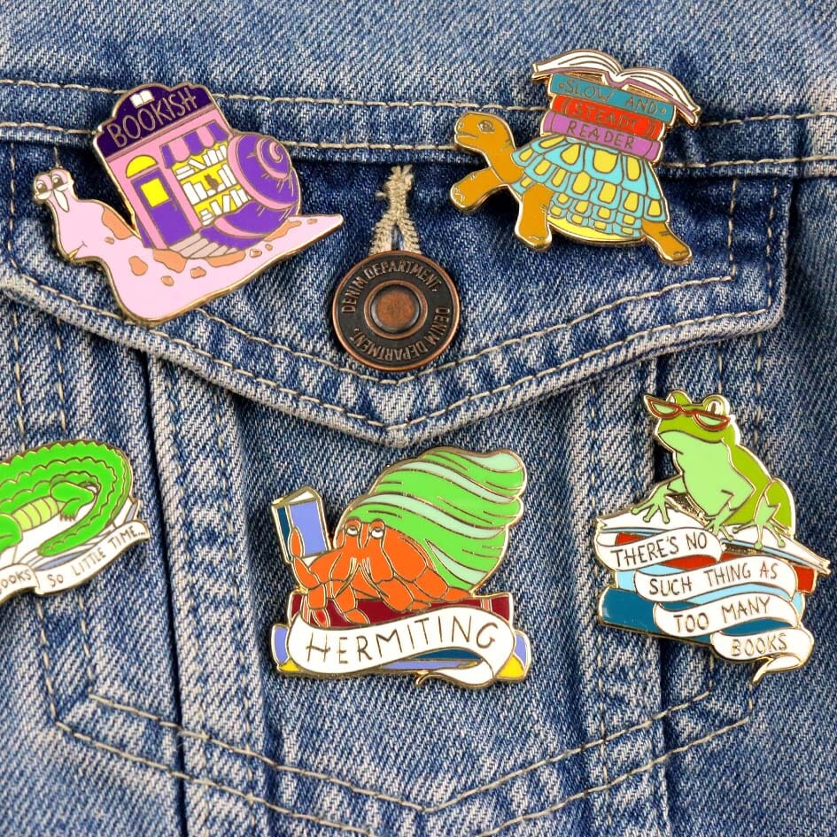 A group of hard enamel pins being displayed on a denim jacket. The pin is in the shape of a Hermit Crab reading a book. The pin says Hermit Crab.