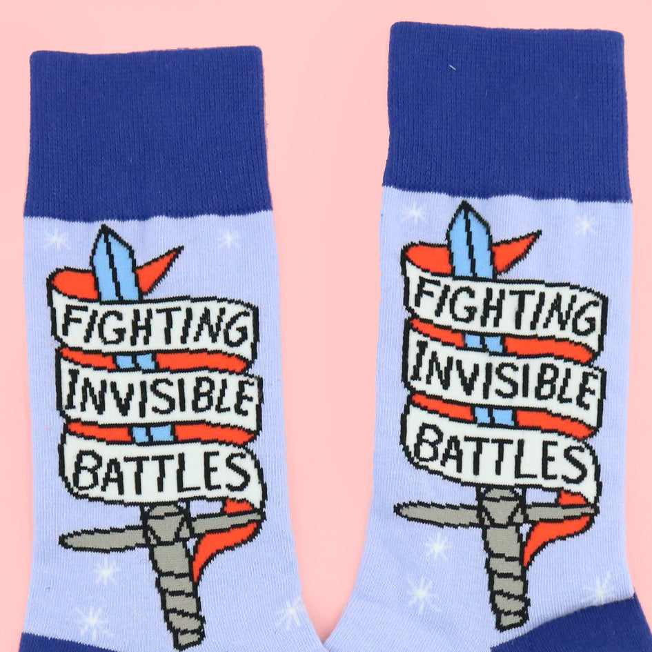 A pair of socks standing against a pink background. The socks are blue and read Fighting Invisible Battles inside a dagger.