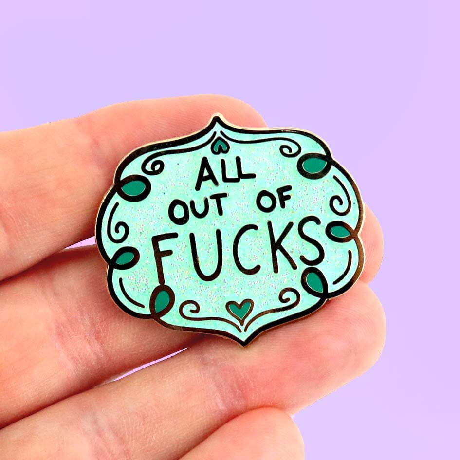 A hard enamel lapel pin being held in a hand against a purple background. The pin has blue sparkly enamel and reads All Out Of Fucks.
