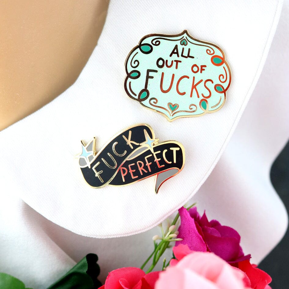 Two hard enamel lapel pins being worn on a white collar. The first pin has blue sparkly enamel and reads All Out Of Fucks. The second pin is black and gray enamel and reads Fuck Perfect.