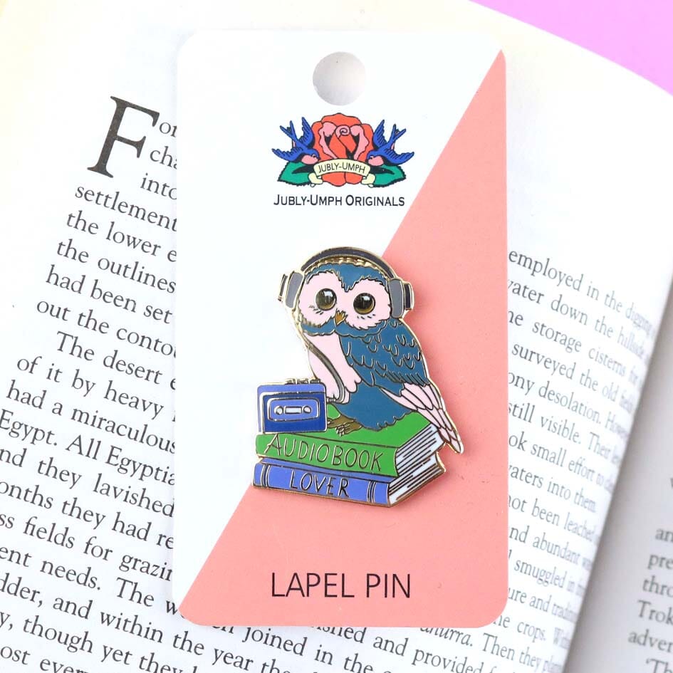 A hard enamel lapel pin on Jubly-Umph cardstock. The pin is in the shape of an owl sitting on a stack of books wearing headphones. The pin reads Audiobook Lover.