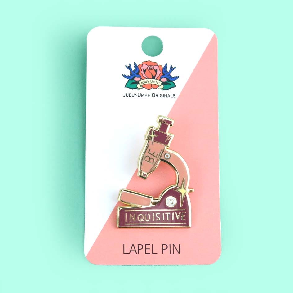 A hard enamel lapel pin on Jubly-Umph cardstock. The pin is in the shape of a microscope and reads Be Inquisitive.