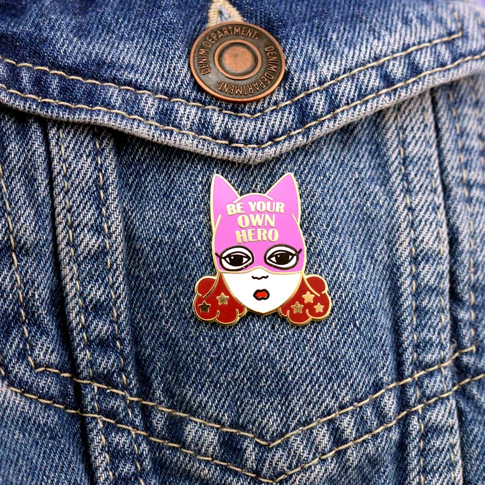 A hard enamel lapel pin against a denim jacket. The pin is a white woman with brown hair and a superhero hat that says Be Your Own Hero.