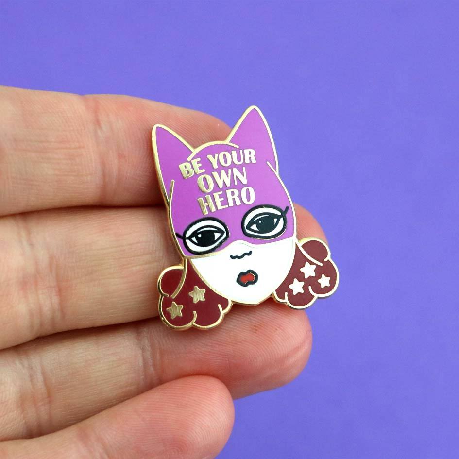 A hard enamel lapel pin being held in the hand. The pin is a white woman with brown hair and a superhero hat that says Be Your Own Hero.