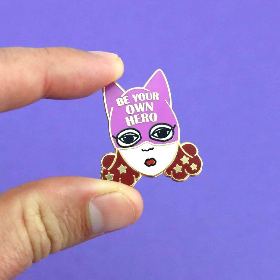 A hard enamel lapel pin being held in the hand. The pin is a white woman with brown hair and a superhero hat that says Be Your Own Hero.