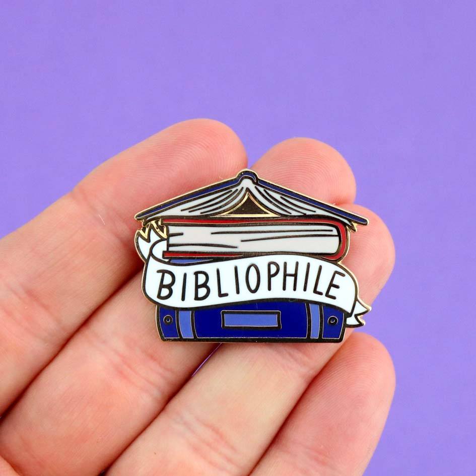 A hard enamel lapel pin being held in a hand against a purple background. The pin says Bibliophile in the middle of a stack of blue books.