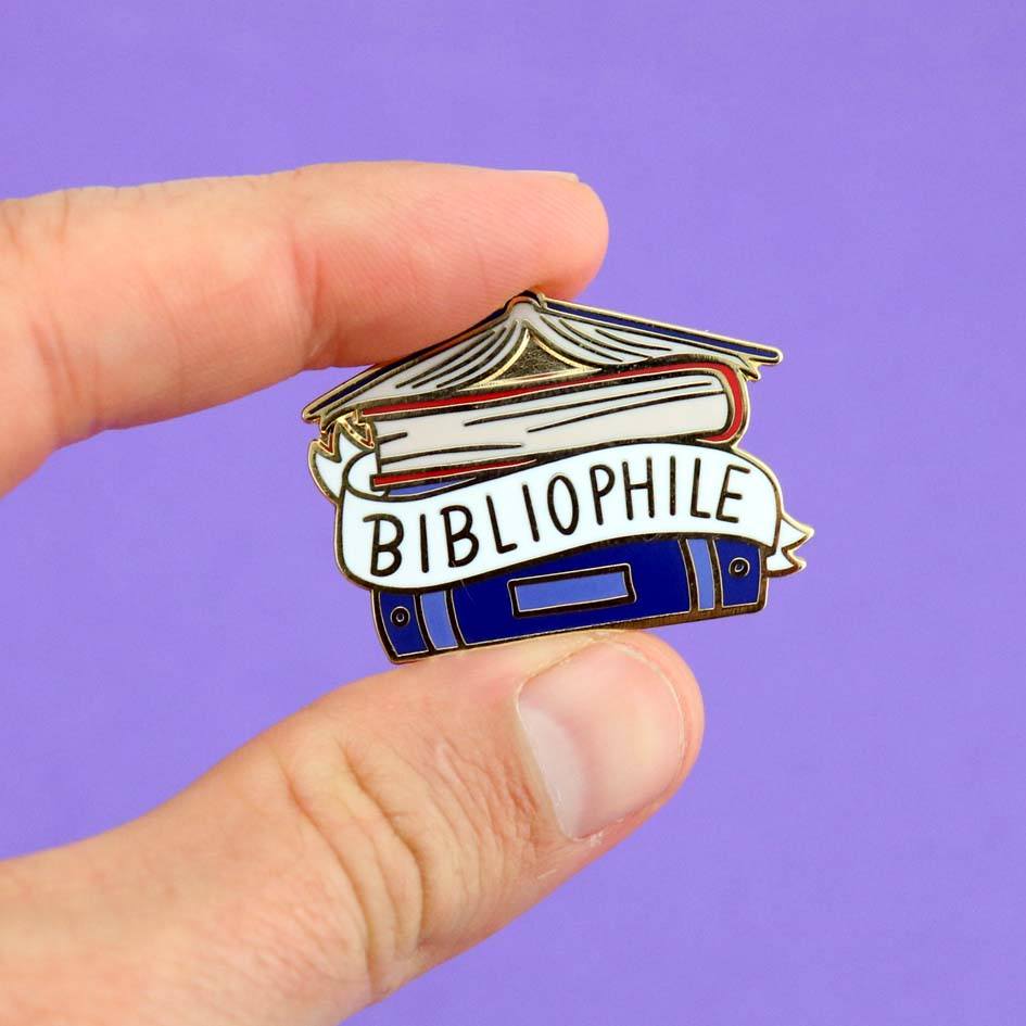 A hard enamel lapel pin being held in a hand against a purple background. The pin says Bibliophile in the middle of a stack of blue books.
