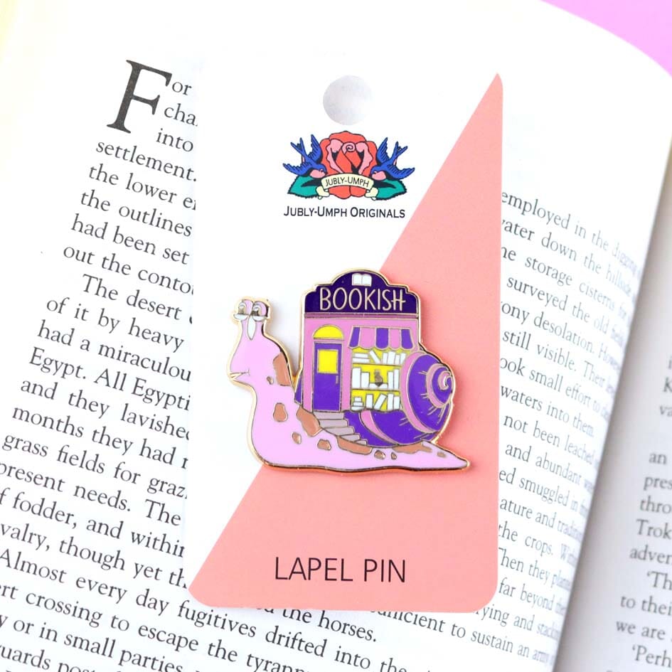 A hard enamel lapel pin on Jubly-Umph cardstock. The pin says say Bookish. The pin design is a snail with a bookshop on its back.