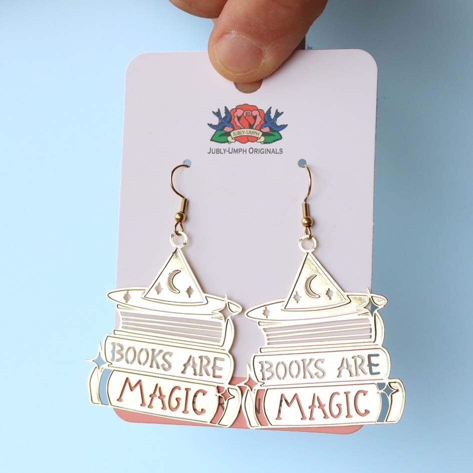 A pair of dangle brass earrings displayed on Jubly-Umph cardstock. The earrings say Books are Magic with books and a witch’s hat.  