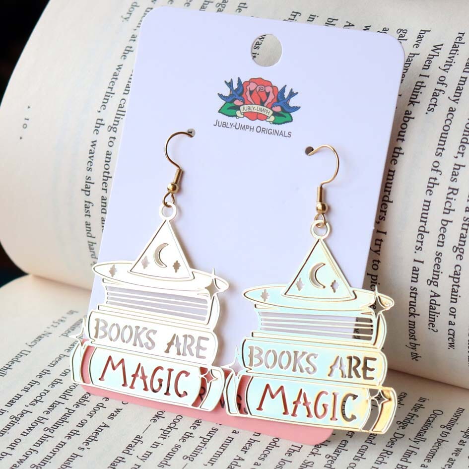 A pair of dangle brass earrings displayed on Jubly-Umph cardstock. The earrings say Books are Magic with books and a witch’s hat.  