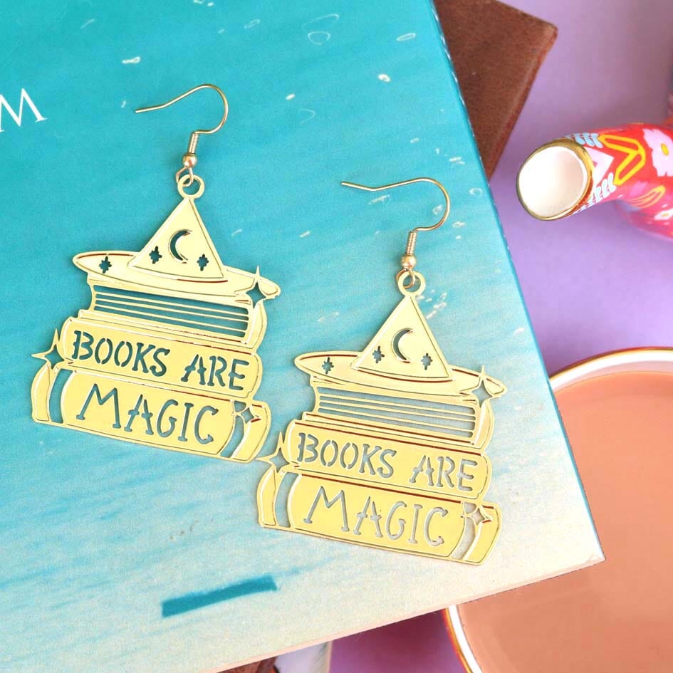 A pair of dangle brass earrings displayed on a blue book background. The earrings say Books are Magic with books and a witch’s hat.  