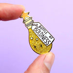 A hard enamel lapel pin being held in a hand. The pin is in the profile of a bottle with yellow glitter. The pin says Bottle of Boldness.