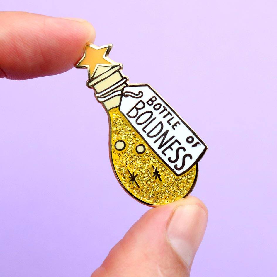 A hard enamel lapel pin being held in a hand. The pin is in the profile of a bottle with yellow glitter. The pin says Bottle of Boldness.
