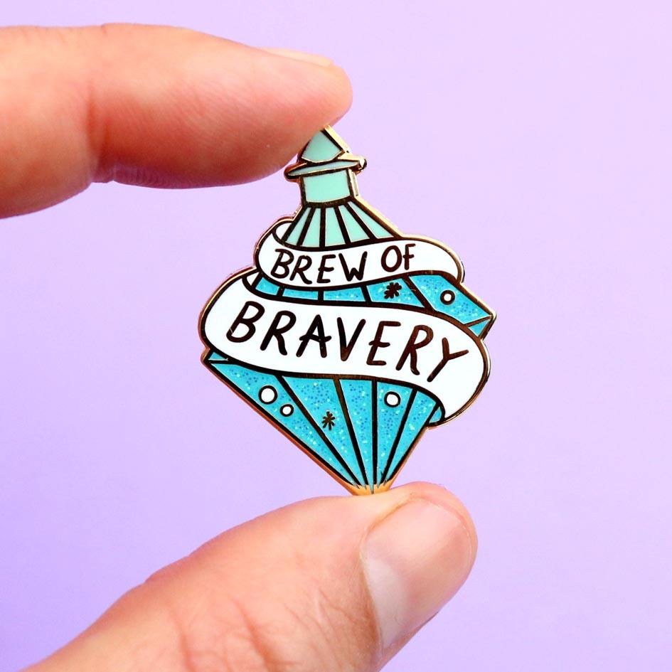 A hard enamel lapel pin being held in a hand. The pin is in the profile of a diamond shaped bottle with blue glitter. The pin says Brew Of Bravery.