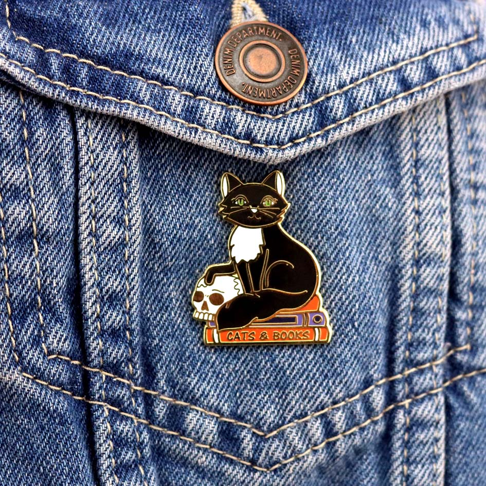 A hard enamel lapel pin against a denim jacket. The pin is in the shape of a black and white cat with a scull, sitting on books. The pin reads Cat’s and Books.